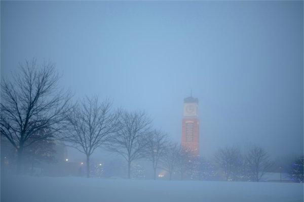  黎明时分，雾气弥漫的大学校园里，一排树木后面是一座钟楼. 