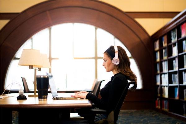  一名大学生戴着pick耳机在大学图书馆的笔记本电脑上工作. 