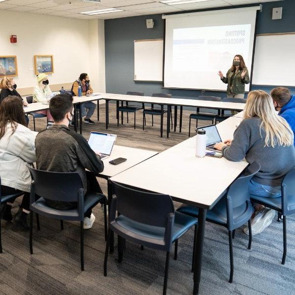 Jessica Fillmore leads a small group workshop, standing and speaking at the front of the room, students in chairs at tables shaped like a box. Screen reads Spotting Procrastination